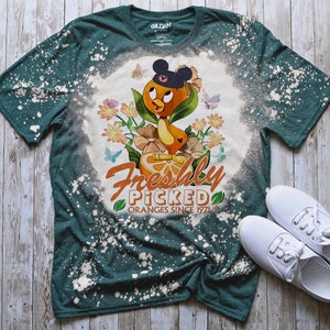 May include: A teal blue t-shirt with a bleach-splattered design featuring a cartoon orange bird wearing a Mickey Mouse hat and holding an orange. The text "Freshly Picked Oranges Since 1971" is printed below the bird.