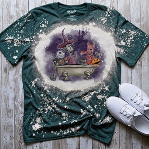 K&ouml;nnte beinhalten: Ein gr&uuml;nes T-Shirt mit einem bleichmittelgesprenkelten Design, das Figuren aus dem Film Nightmare Before Christmas zeigt. Die Figuren befinden sich in einer Badewanne mit K&uuml;rbissen und einem K&auml;fig. Das Shirt hat ein Gildan-Etikett.