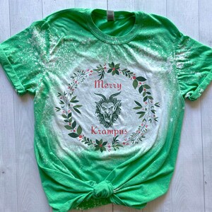 May include: Green t-shirt with a bleach-splattered design featuring a wreath with the text "Merry Krampus" and a cartoon Krampus inside the wreath.