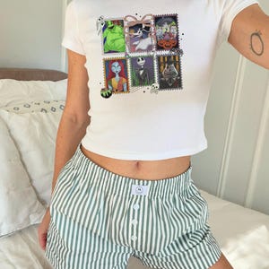 May include: White t-shirt featuring a graphic of six postage stamps with characters from The Nightmare Before Christmas. The shirt is worn with green and white striped boxer shorts. The shorts have a button closure.