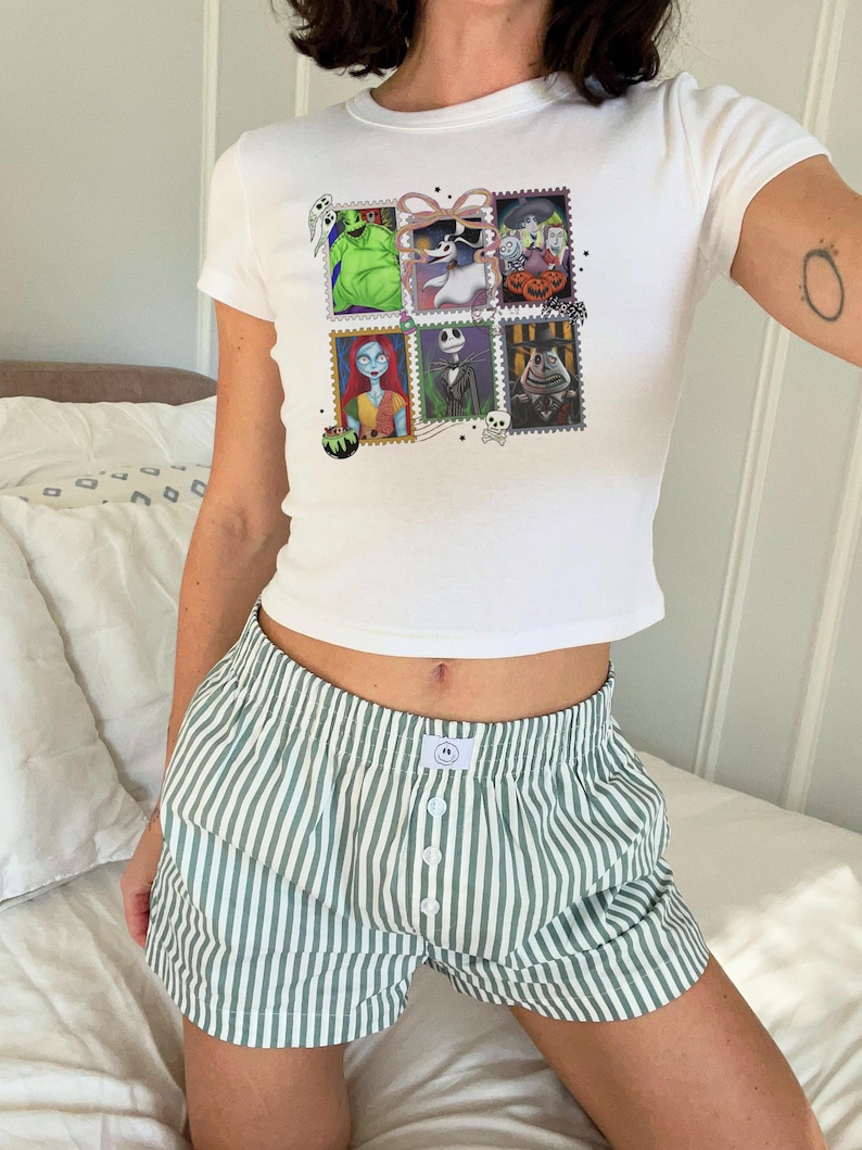 May include: White t-shirt featuring a graphic of six postage stamps with characters from The Nightmare Before Christmas. The shirt is worn with green and white striped boxer shorts. The shorts have a button closure.