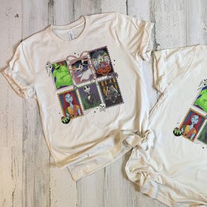 May include: Two white t-shirts with a colourful graphic featuring characters from The Nightmare Before Christmas. The design is arranged in a stamp-like format, showcasing Jack Skellington, Sally, and other characters. The shirts are made of a soft material.