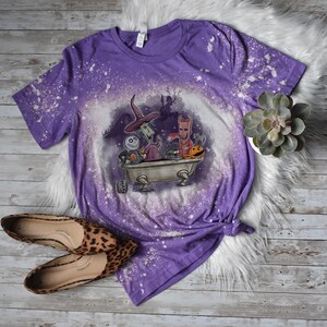 K&ouml;nnte beinhalten: Lilafarbenes T-Shirt mit einem Bleichmittel-Spritzmuster, das Figuren aus dem Film Nightmare Before Christmas zeigt. Die Figuren befinden sich in einer Badewanne mit einem K&uuml;rbis und anderen Halloween-Dekorationen.