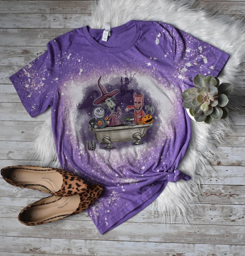K&ouml;nnte beinhalten: Lilafarbenes T-Shirt mit einem Bleichmittel-Spritzmuster, das Figuren aus dem Film Nightmare Before Christmas zeigt. Die Figuren befinden sich in einer Badewanne mit einem K&uuml;rbis und anderen Halloween-Dekorationen.