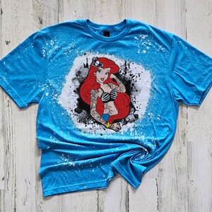 May include: A blue t-shirt with a white bleach design of a red-haired mermaid with a skull and crossbones tattoo. The design is surrounded by black ink splatters.