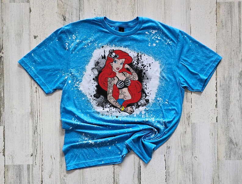 May include: A blue t-shirt with a white bleach design of a red-haired mermaid with a skull and crossbones tattoo. The design is surrounded by black ink splatters.
