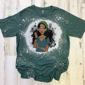 May include: A green t-shirt with a bleach-splattered design featuring a cartoon illustration of a woman with dark skin, green hair, and tattoos. The woman is wearing a green hat and a blue shirt.