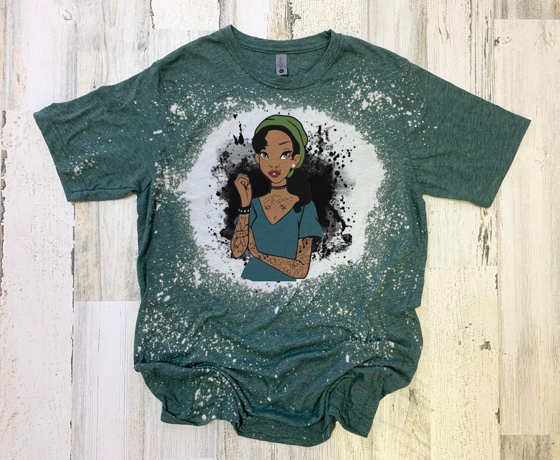 May include: A green t-shirt with a bleach-splattered design featuring a cartoon illustration of a woman with dark skin, green hair, and tattoos. The woman is wearing a green hat and a blue shirt.