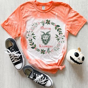 May include: Orange tie-dye t-shirt with a white design of a wreath with a green Krampus inside. The text "Merry Krampus" is written in red inside the wreath.