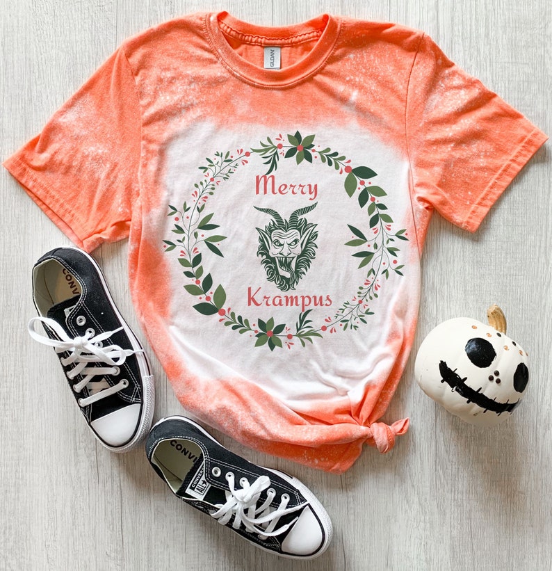 May include: Orange tie-dye t-shirt with a white design of a wreath with a green Krampus inside. The text "Merry Krampus" is written in red inside the wreath.