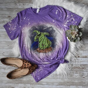 Op de afbeelding: Paars T-shirt met een ontwerp met bleekmiddelspatten, waarop een groen personage uit The Nightmare Before Christmas te zien is. Het personage danst op een platform met spikes.