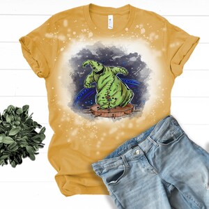 Op de afbeelding: Een geel T-shirt met een ontwerp met bleekmiddelspatten, waarop een groen cartoonfiguur met een gestikt lichaam te zien is. Het figuur staat op een platform met spikes. De achtergrond is een donkerblauwe en paarse galaxie met sterren.