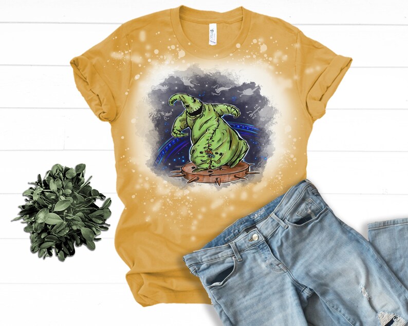 Op de afbeelding: Een geel T-shirt met een ontwerp met bleekmiddelspatten, waarop een groen cartoonfiguur met een gestikt lichaam te zien is. Het figuur staat op een platform met spikes. De achtergrond is een donkerblauwe en paarse galaxie met sterren.