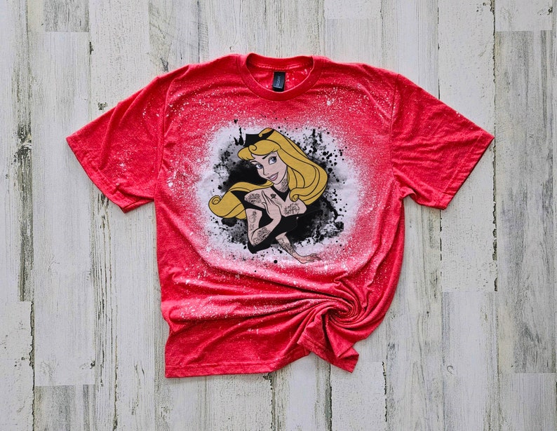 May include: Red t-shirt with a white bleach design of a cartoon princess with blonde hair and a crown. The princess is wearing a black dress and has tattoos on her arms.