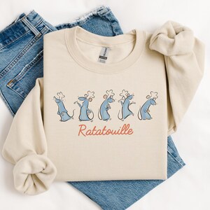 May include: Cream-coloured crew neck sweatshirt featuring a cartoon design of five blue chef rats and the word "Ratatouille" in red. The sweatshirt is folded on blue denim jeans.