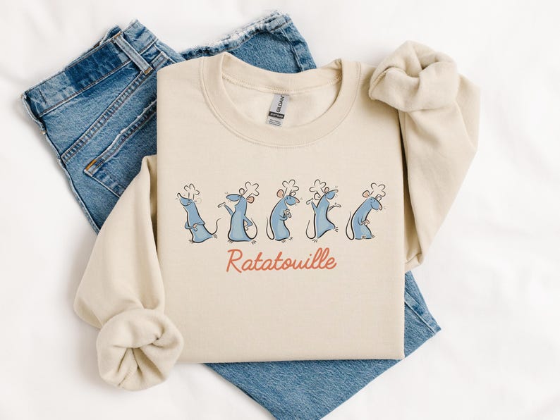 May include: Cream-coloured crew neck sweatshirt featuring a cartoon design of five blue chef rats and the word "Ratatouille" in red. The sweatshirt is folded on blue denim jeans.