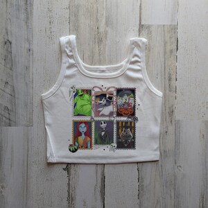 May include: White vest top with a colourful graphic of postage stamp-style images of characters from The Nightmare Before Christmas. The images include Jack Skellington, Sally, and Oogie Boogie. The vest top has a scoop neck and wide straps.