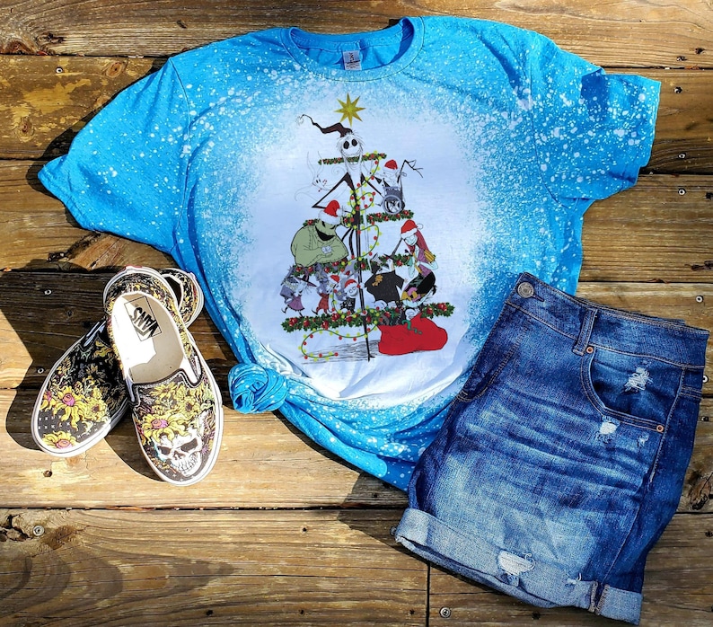 May include: A blue tie-dye t-shirt with a white Christmas tree design featuring characters from the Nightmare Before Christmas. The tree is decorated with ornaments and lights. The shirt is paired with denim shorts and black and yellow floral Vans shoes.