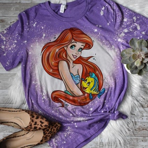May include: Purple t-shirt with a bleach splattered design featuring a cartoon image of Ariel from The Little Mermaid holding Flounder.