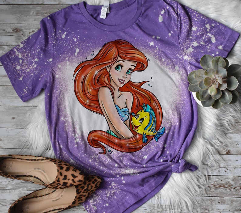 May include: Purple t-shirt with a bleach splattered design featuring a cartoon image of Ariel from The Little Mermaid holding Flounder.