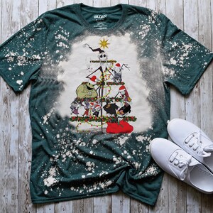 May include: A green t-shirt with a bleach-splattered design featuring a Christmas tree with characters from The Nightmare Before Christmas. The tree is decorated with ornaments and lights, and the characters are all wearing Santa hats.