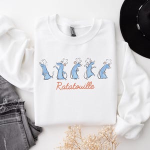 May include: White sweatshirt with a cartoon design of blue rats in chef hats, and the word "Ratatouille" in orange script. The sweatshirt is styled with grey jeans and a black hat.