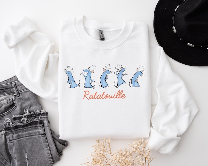 May include: White sweatshirt with a cartoon design of blue rats in chef hats, and the word "Ratatouille" in orange script. The sweatshirt is styled with grey jeans and a black hat.