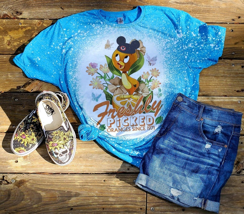 May include: A blue t-shirt with a bleach-splattered design featuring a cartoon duck wearing a Mickey Mouse hat and holding an orange. The text "Freshly Picked Oranges Since 1971" is printed below the duck.