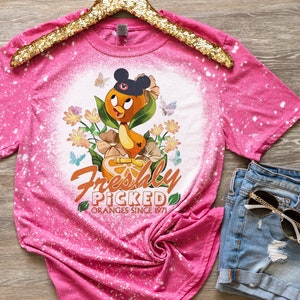 May include: Pink bleached t-shirt with a cartoon orange bird wearing a Mickey Mouse hat and holding an orange. The shirt has the text "Freshly Picked Oranges Since 1971" printed on it.