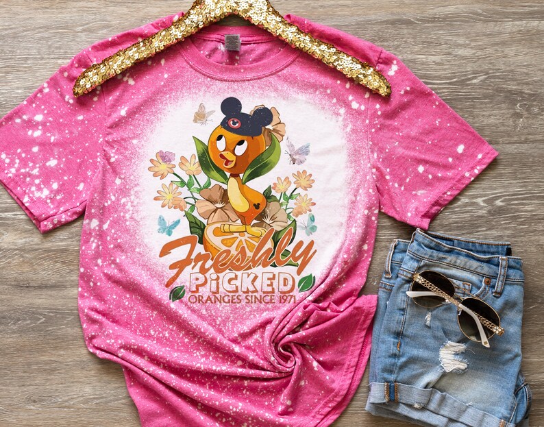 May include: Pink bleached t-shirt with a cartoon orange bird wearing a Mickey Mouse hat and holding an orange. The shirt has the text "Freshly Picked Oranges Since 1971" printed on it.