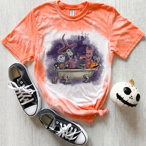 Pode incluir: Camiseta laranja tie-dye com efeito de respingos de lixívia. A camiseta apresenta um gráfico de personagens do filme O Estranho Mundo de Jack em uma banheira. Os personagens incluem Jack Skellington, Sally e Oogie Boogie.