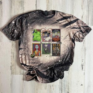 May include: A short-sleeved t-shirt with a black and white speckled pattern. The shirt features six postage stamp-style images of characters from The Nightmare Before Christmas. The images include Jack Skellington, Sally, and Oogie Boogie.