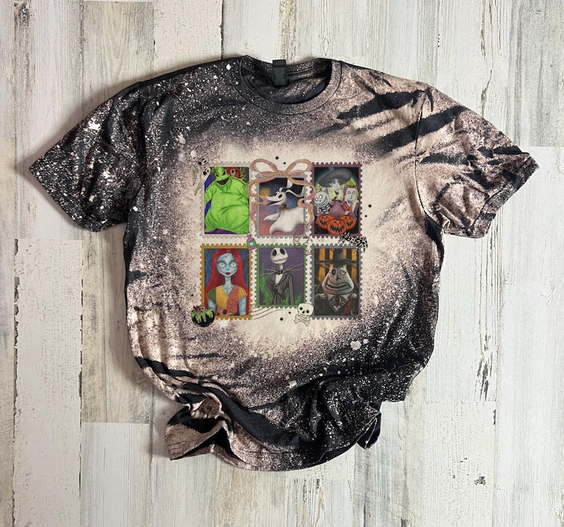 May include: A short-sleeved t-shirt with a black and white speckled pattern. The shirt features six postage stamp-style images of characters from The Nightmare Before Christmas. The images include Jack Skellington, Sally, and Oogie Boogie.