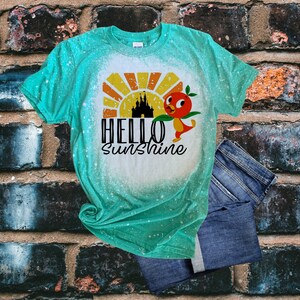 May include: A turquoise blue t-shirt with a bleach-splattered design. The shirt features a cartoon bird with a red beak and orange body, a sunset, and a castle silhouette. The text "HELLO SUNSHINE" is printed in black.