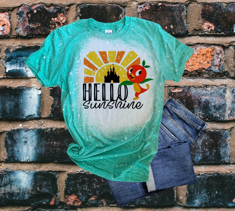 May include: A turquoise blue t-shirt with a bleach-splattered design. The shirt features a cartoon bird with a red beak and orange body, a sunset, and a castle silhouette. The text "HELLO SUNSHINE" is printed in black.