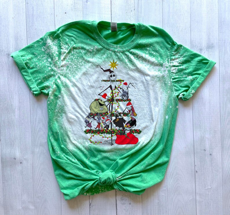 May include: Green t-shirt with a bleach-splattered design featuring a Christmas tree with characters from the Nightmare Before Christmas. The tree is decorated with lights, ornaments, and a star on top. The characters include Jack Skellington, Sally, and Oogie Boogie.