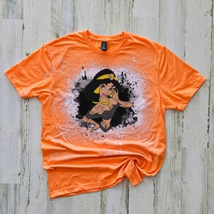 May include: Orange t-shirt with a bleach-splattered design featuring a cartoon image of Princess Jasmine from Aladdin. The design is in black, white, and yellow.