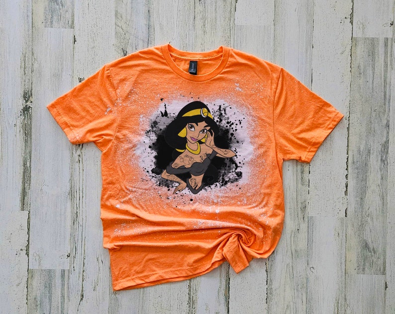 May include: Orange t-shirt with a bleach-splattered design featuring a cartoon image of Princess Jasmine from Aladdin. The design is in black, white, and yellow.