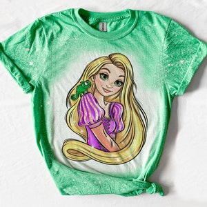 May include: Green t-shirt with a bleach-splattered design featuring a cartoon image of Rapunzel from Disney's Tangled. Rapunzel is wearing a purple dress and has long blonde hair. A green chameleon is on her shoulder.