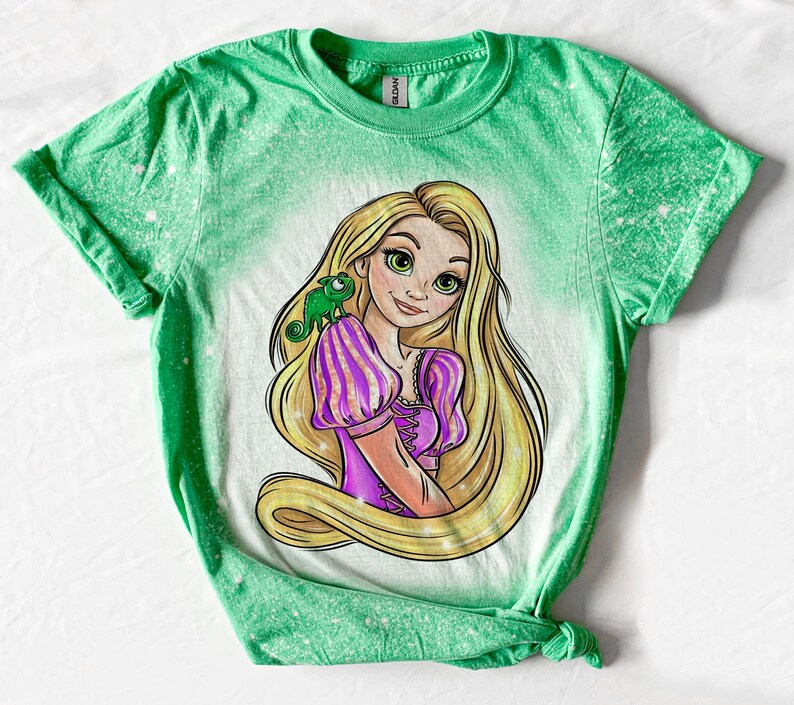 May include: Green t-shirt with a bleach-splattered design featuring a cartoon image of Rapunzel from Disney's Tangled. Rapunzel is wearing a purple dress and has long blonde hair. A green chameleon is on her shoulder.
