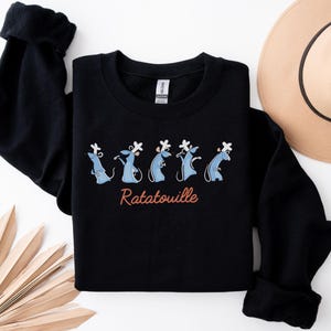 May include: Black sweatshirt with a cartoon design of blue rats wearing chef hats, and the word "Ratatouille" in a cursive font. A tan hat and a decorative fan are also visible.