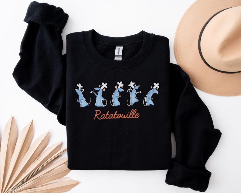 May include: Black sweatshirt with a cartoon design of blue rats wearing chef hats, and the word "Ratatouille" in a cursive font. A tan hat and a decorative fan are also visible.