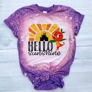May include: Purple tie-dye t-shirt with a bleached design featuring a cartoon bird, a castle, and a sunset. The text "HELLO SUNSHINE" is printed on the shirt.
