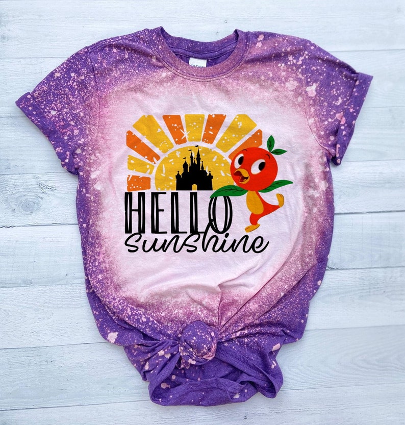 May include: Purple tie-dye t-shirt with a bleached design featuring a cartoon bird, a castle, and a sunset. The text "HELLO SUNSHINE" is printed on the shirt.