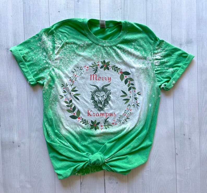 May include: Green t-shirt with a bleach-splattered design featuring a wreath with the text "Merry Krampus" and a cartoon Krampus inside the wreath.