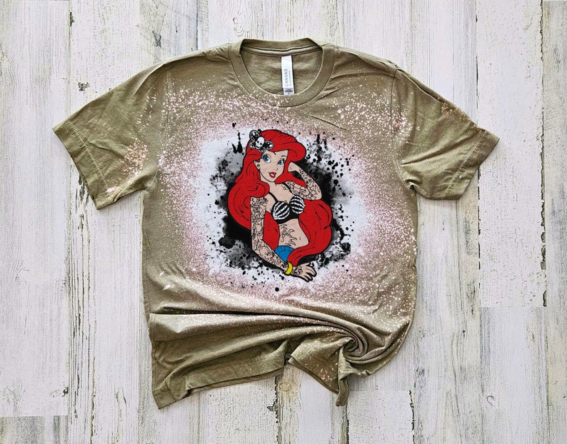 May include: A green t-shirt with a white bleach design of a red-haired mermaid with tattoos. The mermaid is wearing a black bikini top and has a blue tail.