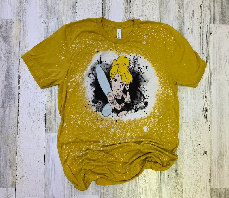 May include: A yellow t-shirt with a bleach-splattered design featuring a cartoon image of Tinkerbell with tattoos. The design is in black and white with a blue fairy wing.