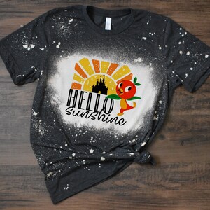 May include: A dark gray t-shirt with a bleach-splattered design. The design features a cartoon bird with a peach, a castle silhouette, and the text "HELLO SUNSHINE".