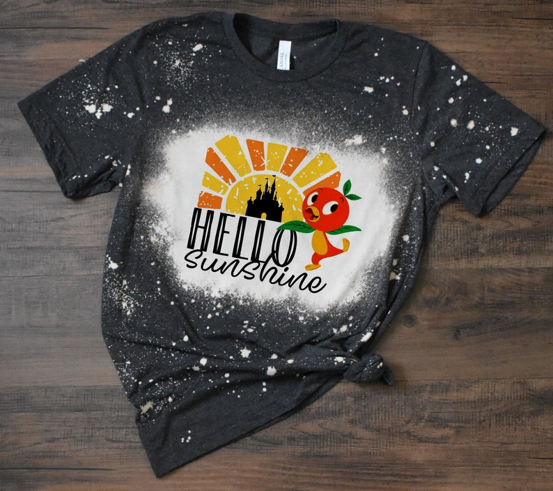 May include: A dark gray t-shirt with a bleach-splattered design. The design features a cartoon bird with a peach, a castle silhouette, and the text "HELLO SUNSHINE".