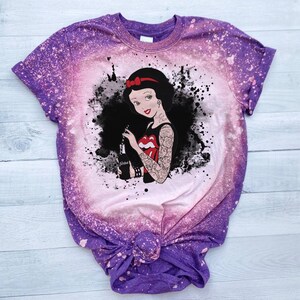 May include: Purple and pink tie-dye t-shirt with a black and white graphic of Snow White with tattoos and holding a bottle of soda. The graphic has a splatter effect.